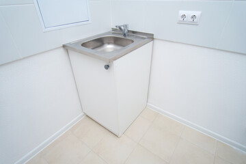 Empty stainless steel sink with faucet in a simple white room. Minimalist utility space with tiled floor and power outlet.