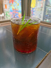 Vertical close-up of a glass of iced cold brew Americano with ice cubes, garnished with two long green leaves, sitting on a dark tiled table with a vibrant, blurred glass window backdrop.