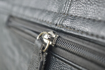close-up of a silver metal zipper pull and teeth on dark gray textured leather. Represents detail of fashion, bag, luggage hardware, industrial, and material texture.