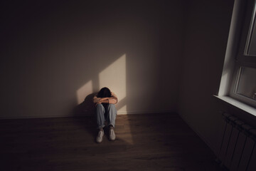 A woman sits alone on the floor in a dimly lit room, hugging their knees, expressing sadness, isolation, and emotional distress.