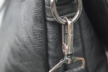 close-up of a silver metal swivel clasp, carabiner clip attached to a dark gray textured leather bag. Represents fashion accessory, hardware detail, luxury, and material texture.