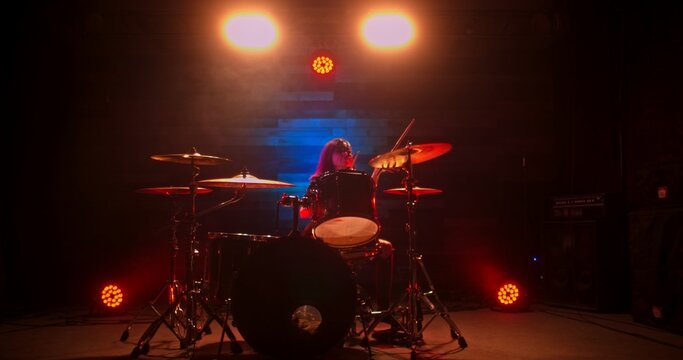 Professional female drummer with red hair playing on stage during rock show in nightclub. Girl vigorously beats the drums with drumsticks.