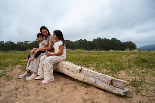 A woman sits on a log with two children. The children are wearing dresses and the woman is holding a cell phone. Scene is calm and peaceful, as the family enjoys their time together on the beach