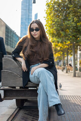 Fototapeta premium Fashion-forward young Asian woman wearing sunglasses, blazer and denim jeans, sitting on an industrial-style bench in a modern city street. Urban lifestyle, street fashion and contemporary vibe