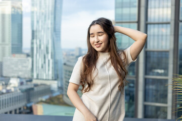 Fototapeta premium Asian woman in chic beige dress holding a handbag on a rooftop terrace with city skyline — full-length fashion & lifestyle portrait