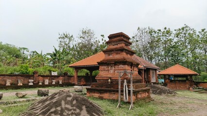 A traditional red-brick temple surrounded by lush greenery and ancient stone relics in a rural Indonesian landscape. This image is perfect for cultural tourism, historical education, and heritage pres