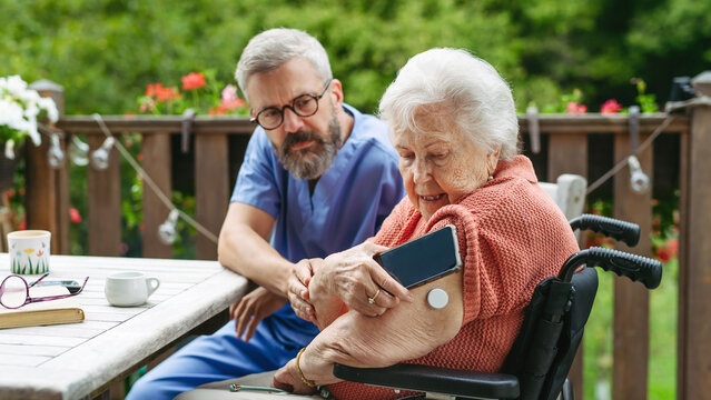 Home nurse checking glucose level of senior lady with smartphone.