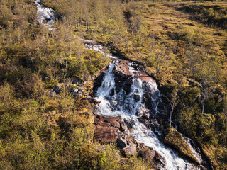 Aerial view of a stunning mountain waterfall cascading through a vibrant green birch forest in Norway.