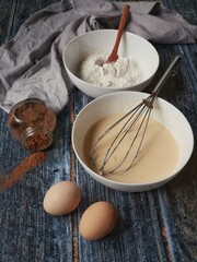 Ingredients for baking on a rustic wooden table - bowls with flour and batter, eggs, cocoa powder, and a whisk.