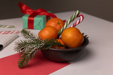 A bowl filled with fresh oranges sits on a table, surrounded by candy canes and a small wrapped gift.