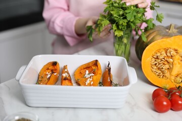 Woman with coriander at white marble table indoors, focus on baked pumpkin