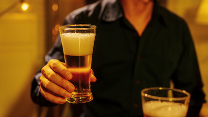 A man in a dark shirt holding a glass of freshly poured beer, golden foam rising under warm lights, symbolizing relaxation, confidence, and social enjoyment in a friendly atmosphere.