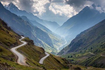 Naklejka premium Winding mountain road descends into a breathtaking valley surrounded by majestic peaks and shrouded in atmospheric clouds during the afternoon light