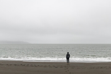 A lone individual man stands on a isolated beach, back turned and looking out in contemplation at a vast fog-enshrouded ocean.