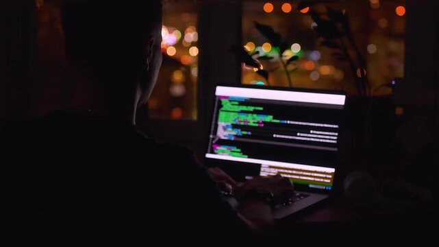 A young male programmer in front of a window with a lights of night city, on his laptop screen is code. out of focus. 