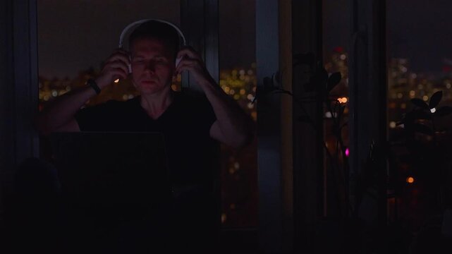 A young male programmer in front of a window with a lights of night city, on his laptop screen is code. out of focus. 