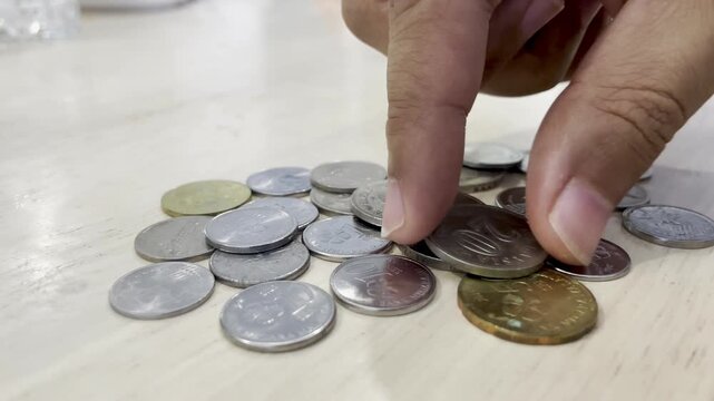 A close-up macro shot shows a hand counting and sorting a scattered pile of Malaysian Ringgit coins on a light wooden table. The shot focuses on finance, savings, and budgeting.