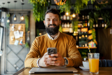 Portrait of smiling business casual man sitting in cafeteria with cellphone in hands and looking at the camera.