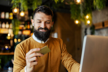 Portrait of a smart casual man sitting in coffee shop with platinum credit card in hands and using it for online shopping.
