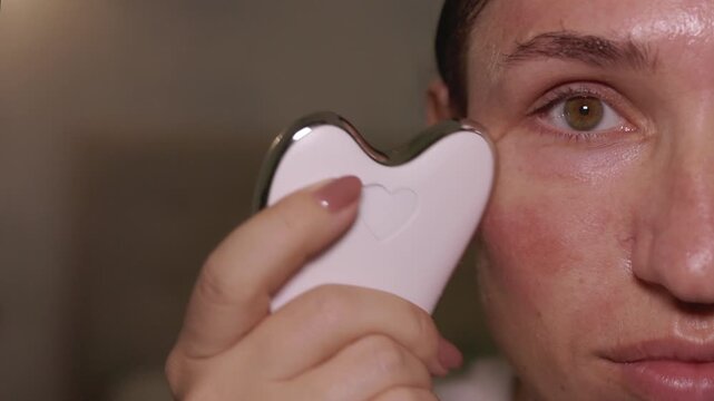 Woman does facial massage with electric gua sha tool with vibration and heat. Close-up portrait of female face, massaging cheeks and forehead. Half of face and eye visible in frame.