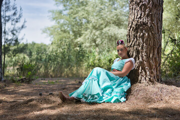 Young woman, beautiful, blonde, with typical Andalusian flamenco costume to go on pilgrimage in light blue color, sitting resting under the shade of a tree. Concept regional costumes, typical, Spanish