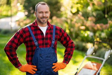 Gardener standing in sunny garden with lawnmower surrounded by green plants