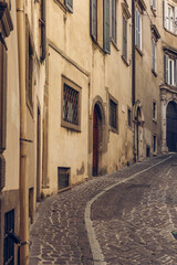 Vintage toned vertical view of a narrow cobblestone street in the Upper Town of Bergamo Italy, curving uphill between aged ochre walls and shuttered windows in warm light