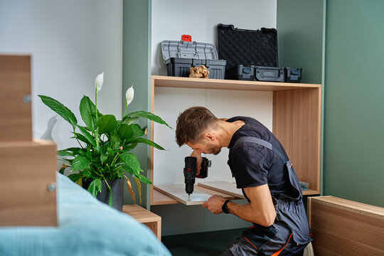 Worker using electric drill to drilling hole in wooden board. Man assembling furniture at home, preparing shelf to installing in self made cabinet. Concept of renovation and interior improvement - Powered by Adobe