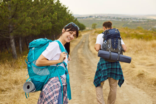 Couple of happy tourists walking on trail by pine forest with backpacks. Young woman turned around to smile while man hiking ahead on dirt road, trekking team adventure and travel in nature of hikers
