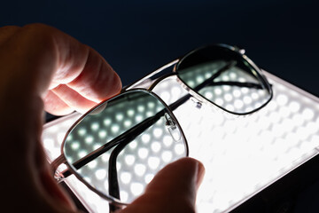 Polished silver frame sunglasses with shaded glass held by hand above bright patterned light panel, contrasted with dramatic black setting in refined fashion presentation