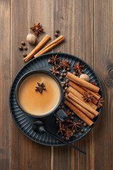 Traditional Indian masala chai tea with milk and spices in a black cup on a dark plate on a wooden background