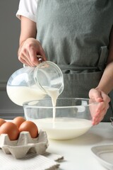 Making batter (liquid dough). Woman pouring milk into bowl at white table indoors, closeup
