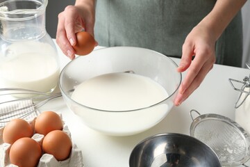 Making batter (liquid dough). Woman adding eggs into bowl at white table indoors, closeup