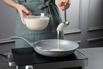 Woman pouring liquid dough from bowl into frying pan at grey table in kitchen, closeup
