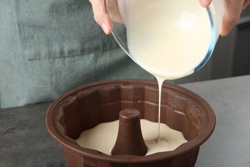 Woman pouring liquid dough from bowl into baking dish at grey table in kitchen, closeup