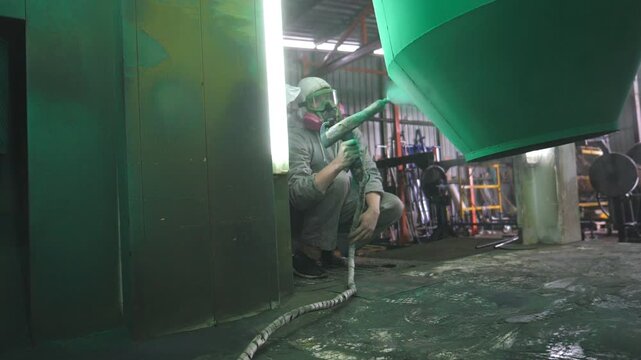 Industrial painting process in chamber at factory. Painter spraying green paint on detail in special booth. Technician in safety wear working at production. Master paint metal in specialised workshop