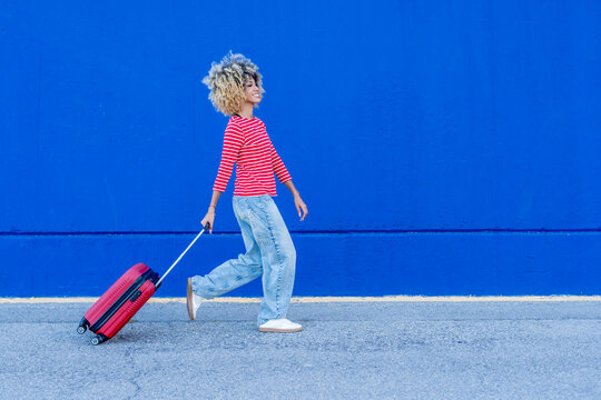 Young woman walking with luggage in front of blue wall