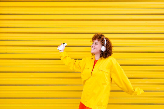 Woman in yellow jacket dancing with smartphone and headphones against a yellow background