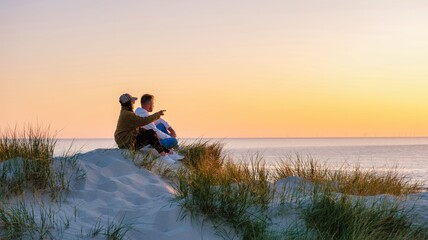 A couple of man and woman sits together on the soft dunes, surrounded by tall grass, gazing at the calm waves of the North Sea during golden hour in Blokhus Strand, Denmark