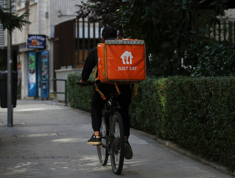 Repartidor rider de Just Eat en bicicleta en ruta y bolsa naranja llevando un pedido de encargo a la direcci&oacute;n de entrega de la comida pagada en l&iacute;nea en plataforma digital con tel&eacute;fono m&oacute;vil app