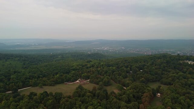 Aerial landscape view from Normafa overlooking the lush green forests of the Buda Hills with the town of Budakeszi visible in the background under cloudy skies.