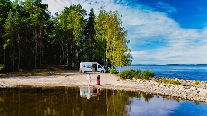 A couple of men and women stand on the tranquil shoreline of Lake Saimaa Finland, surrounded by lush trees and a scenic view. A camper van is parked nearby, inviting adventure and exploration.