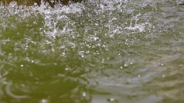 Large drops of water fall from a height into a pond against a rock backdrop. The water, illuminated by the sun, takes on a yellowish color. The falling drops form small dimples and waves on the water.