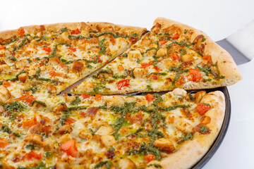 Freshly baked chicken pizza with melted cheese, diced red peppers and drizzled green pesto on thin crust. One slice lifted with metal spatula to show soft interior.