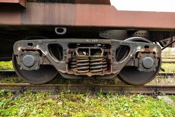 Detailed view of a rusted train bogie showing metal wheels, heavy-duty coil springs, and suspension components over weathered tracks and grassy terrain