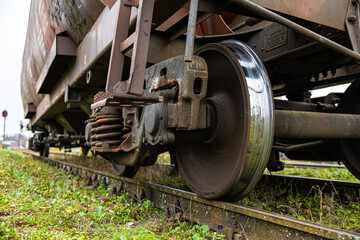 Detailed view of a rusted train bogie showing metal wheels, heavy-duty coil springs, and suspension components over weathered tracks and grassy terrain