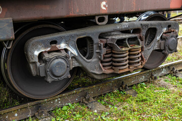 Detailed view of a rusted train bogie showing metal wheels, heavy-duty coil springs, and suspension components over weathered tracks and grassy terrain