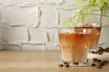 Tasty iced coffee with milk in glasses and beans on wooden table against light textured background, closeup. Space for text