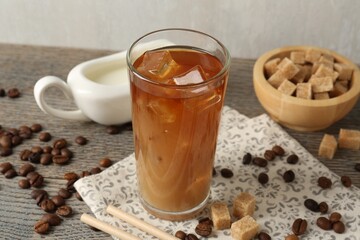 Tasty iced coffee with milk in glass, beans and sugar cubes on wooden table against grey background, closeup