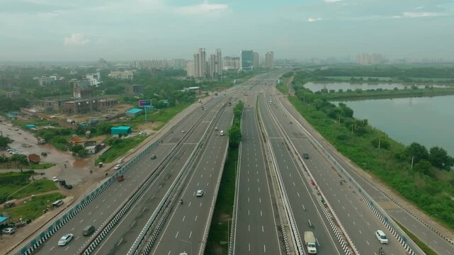 Aerial view of the multi-lane Dwarka Expressway, flanked by lush greenery and a serene lake under a hazy sky, Gurugram, Haryana, India.
